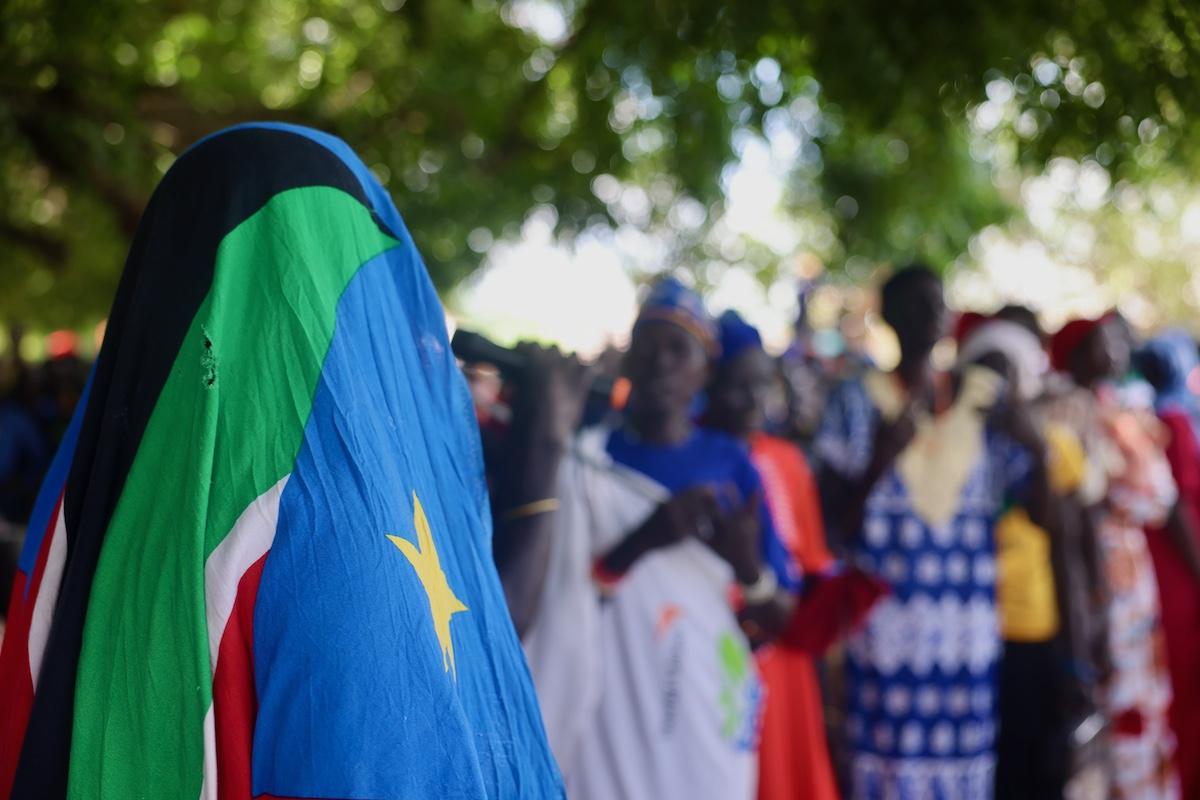 A person in the foreground is facing away from the camera and has covered their head with the South Sudanese flag. In the background, there are more people in bright clothing which have been blurred. All of the these people are standing under trees.