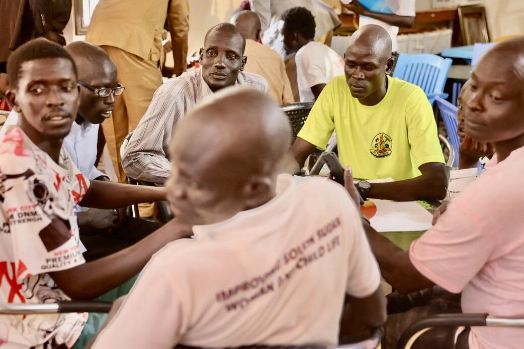 Several people seated around tables indoors, engaged in discussion, with chairs and documents visible in the background.