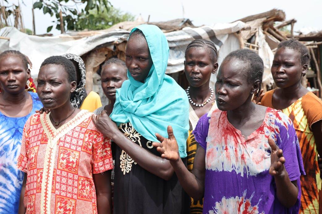 A group of people wearing colorful patterned clothing stand outdoors in front of makeshift shelters, with one person gesturing while others listen.