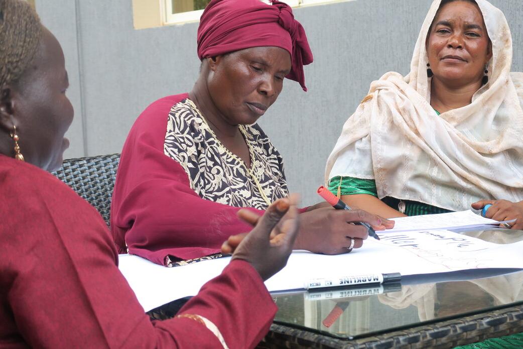A community meeting in a room with orange plastic chairs arranged in rows, participants seated and listening to a speaker at the front near posters and a banner on the wall.