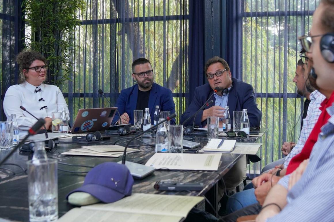 Several individuals seated at a rectangular conference table covered with papers, water bottles, and microphones. A laptop with stickers is visible on the table, along with a blue cap placed near documents. The background shows tall windows with sheer curtains and greenery outside.